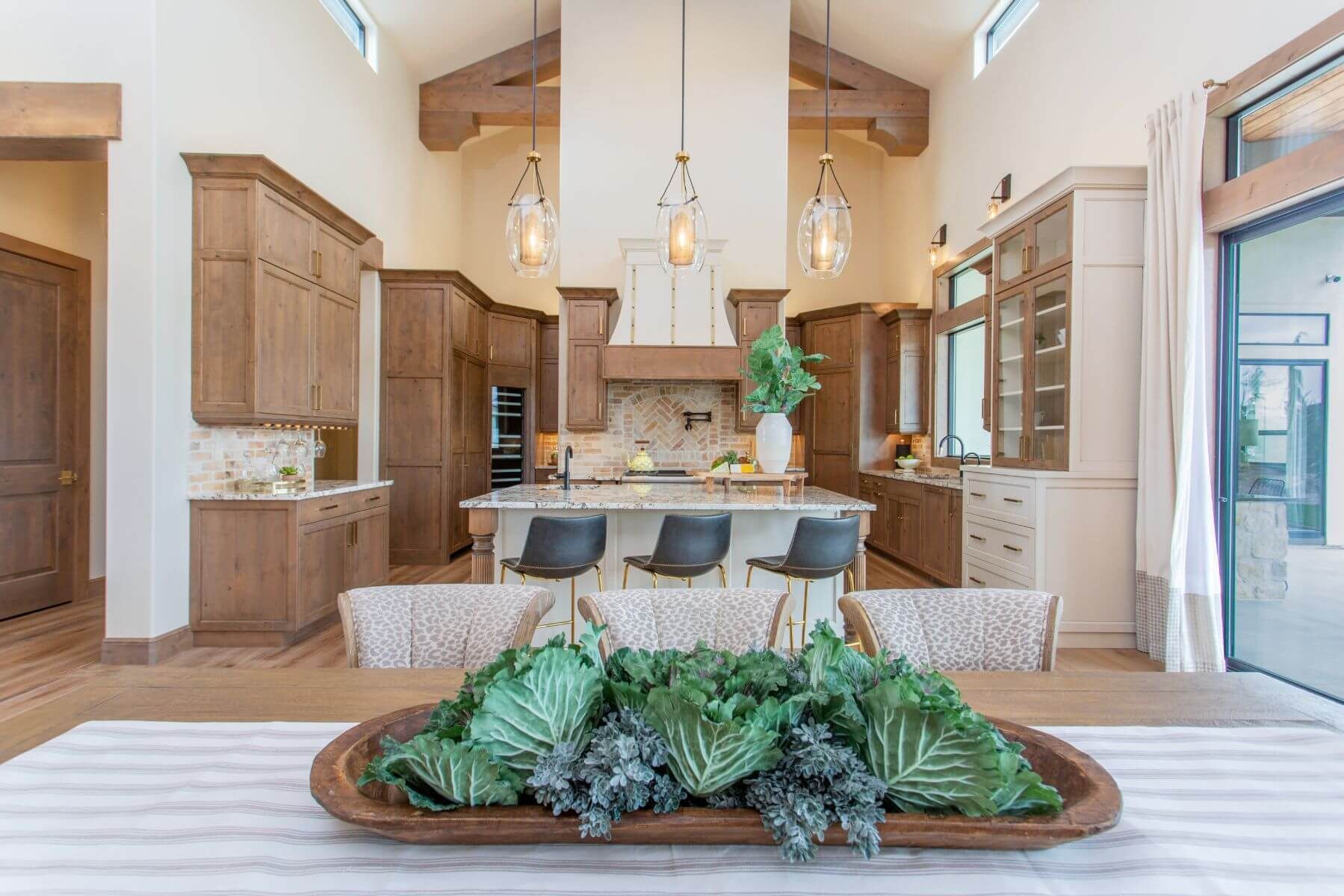 Spacious kitchen with wood cabinets, an island, and decorative lighting, seen from a dining table with a leafy centerpiece.