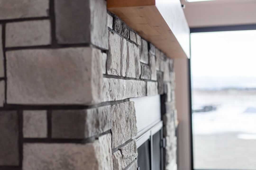 A stone fireplace with a wooden mantle in a living room next to a window.