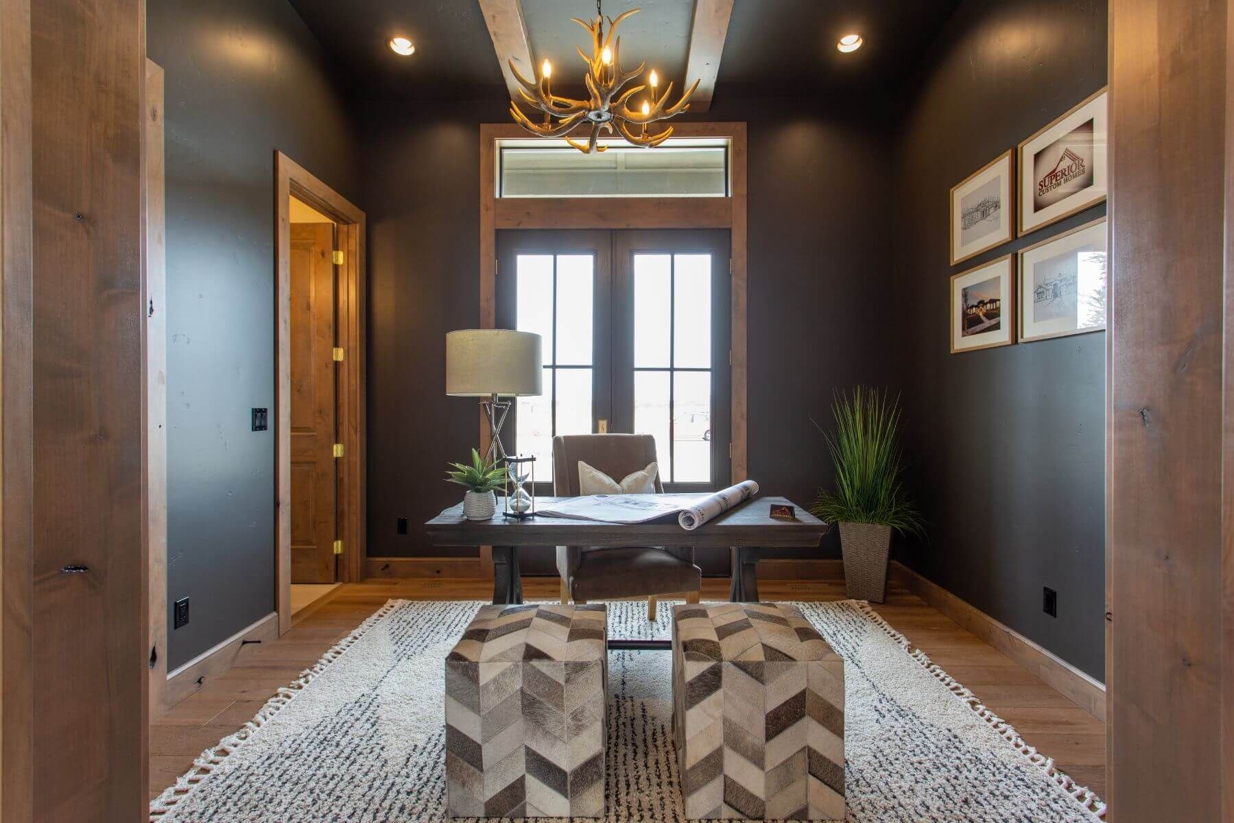 Dark office with wooden trim, desk, and antler chandelier; two patterned ottomans sit on a rug.