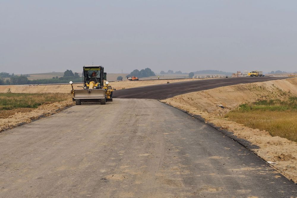 Lavori stradali con livellatrice e asfalto fresco su una strada rurale sotto un cielo fosco.