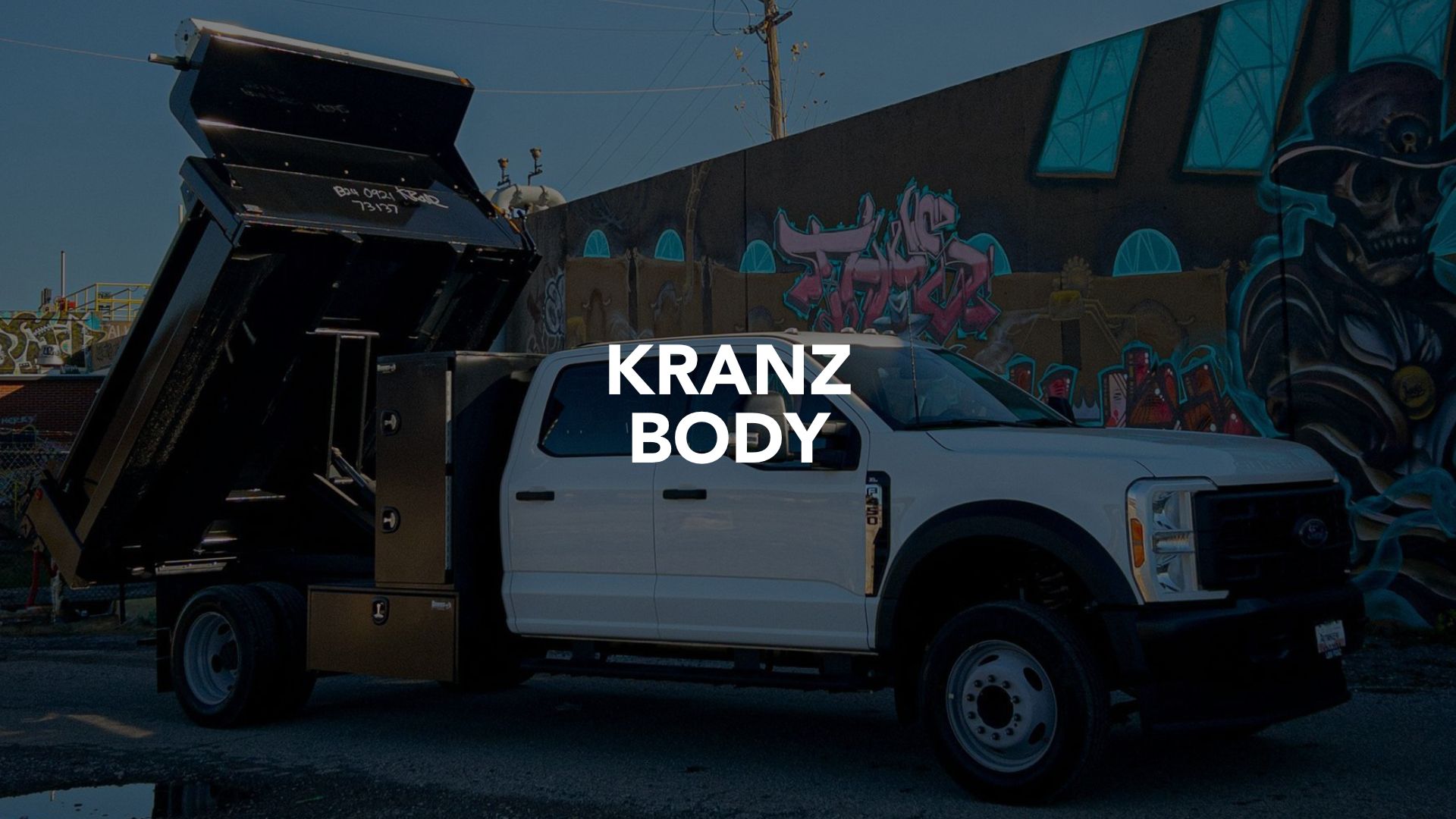 White dump truck with raised bed parked in front of a graffiti wall, overlaid with “KRANZ BODY” text