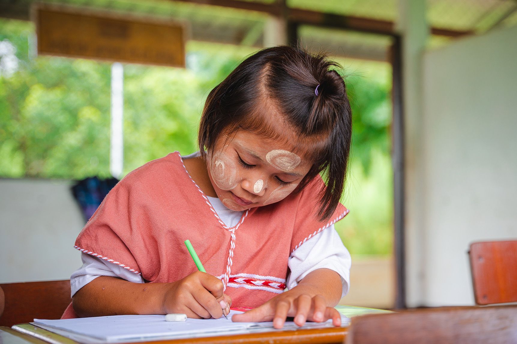 Choose a child to sponsor Young girl with face paint and red shirt, writing at a desk in a classroom.