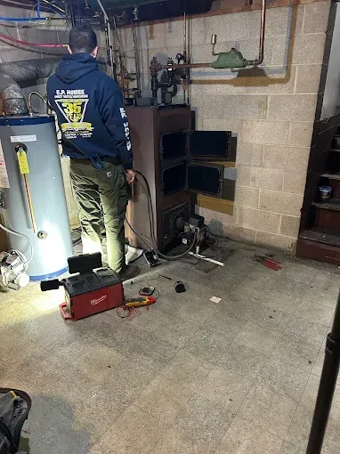 Technician working on a boiler in a basement utility room, with tools and equipment nearby.
