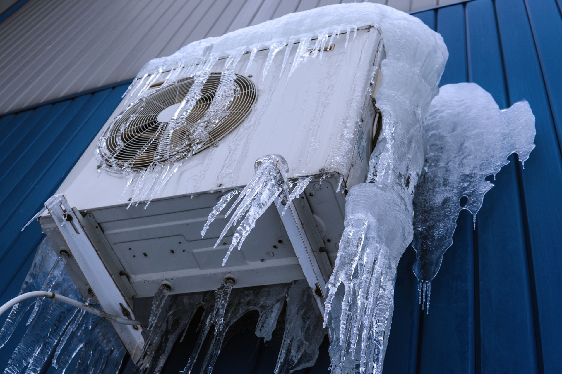 An outdoor HVAC unit mounted on a blue wall is heavily covered in thick ice and long, jagged icicles.