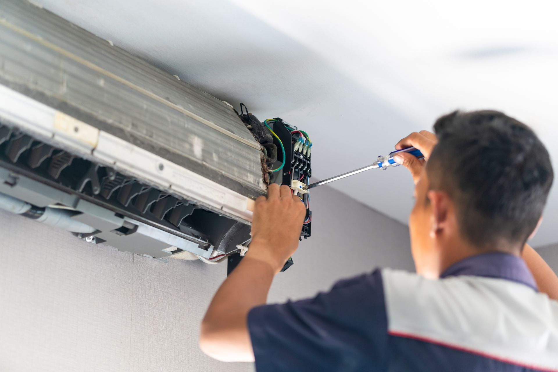 A technician in a two-tone uniform uses a screwdriver to repair the internal electronics of a wall-mounted air conditioner.