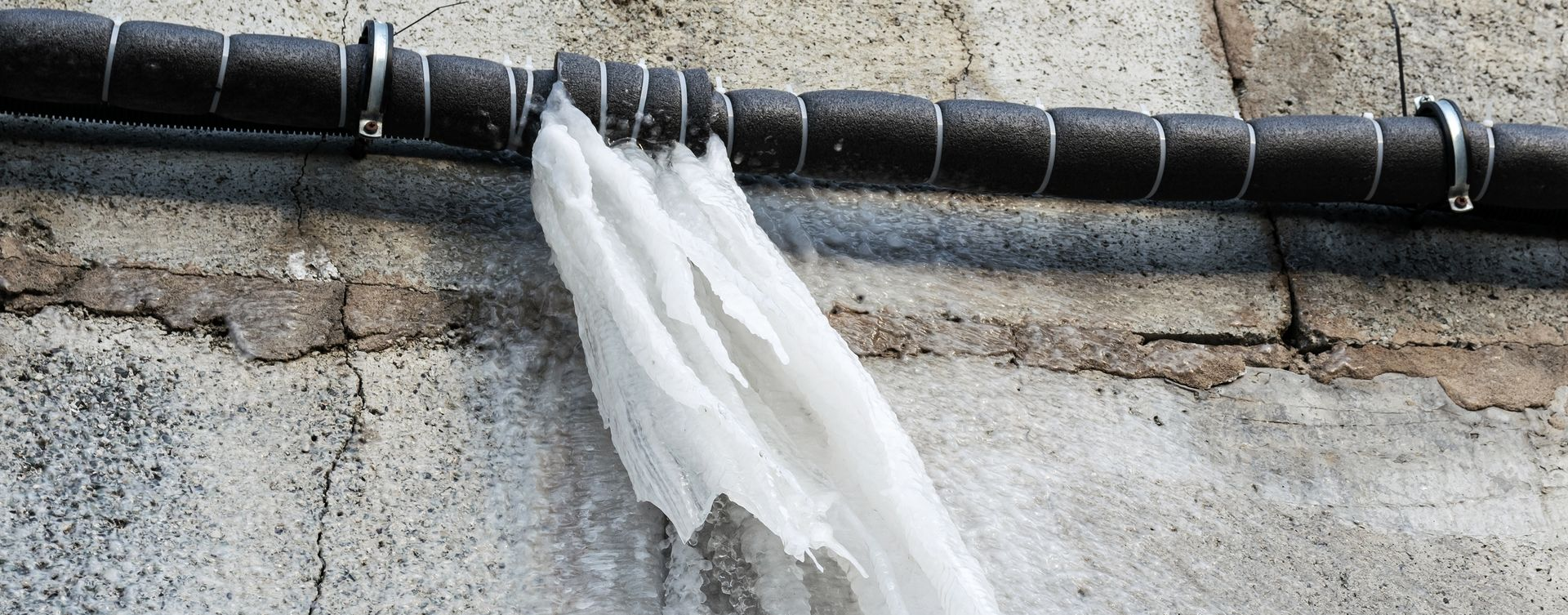 A black, insulated pipe with metal clips mounted on a rough concrete wall, with white shredded plastic hanging from it.