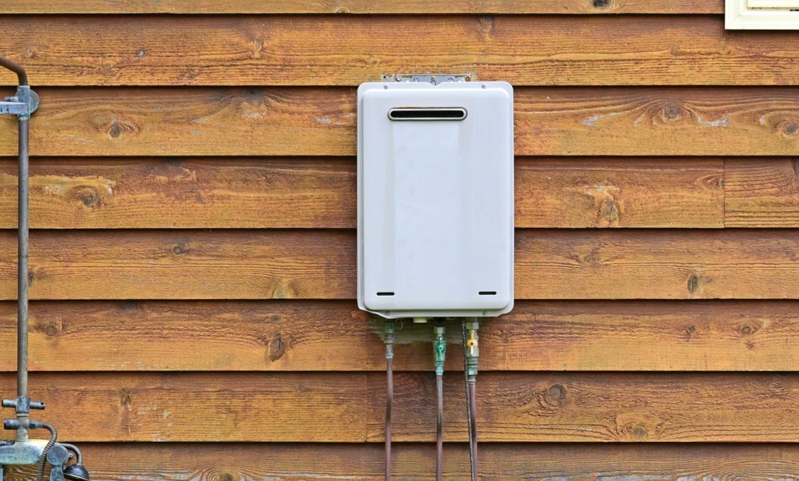 A white, rectangular tankless water heater mounted on a wooden exterior wall with pipes extending from the bottom.