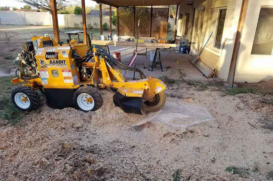 A yellow stump grinder is sitting in the dirt in front of a house.