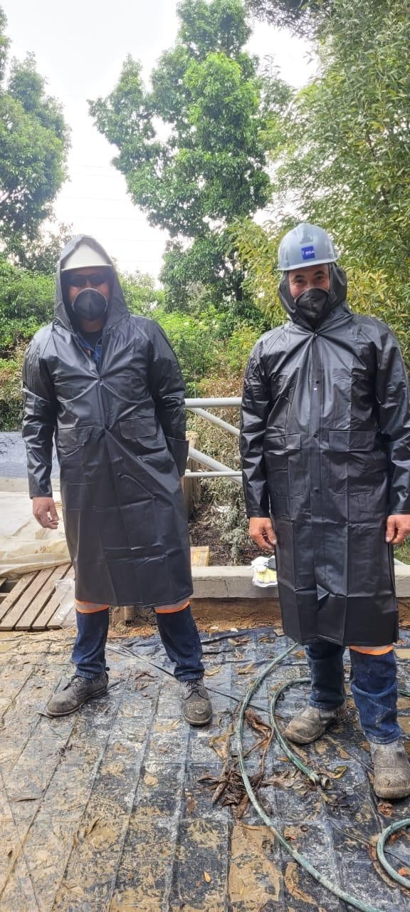 Dos trabajadores con impermeables negros y cascos de seguridad de pie en una obra en construcción mojada bajo los árboles.