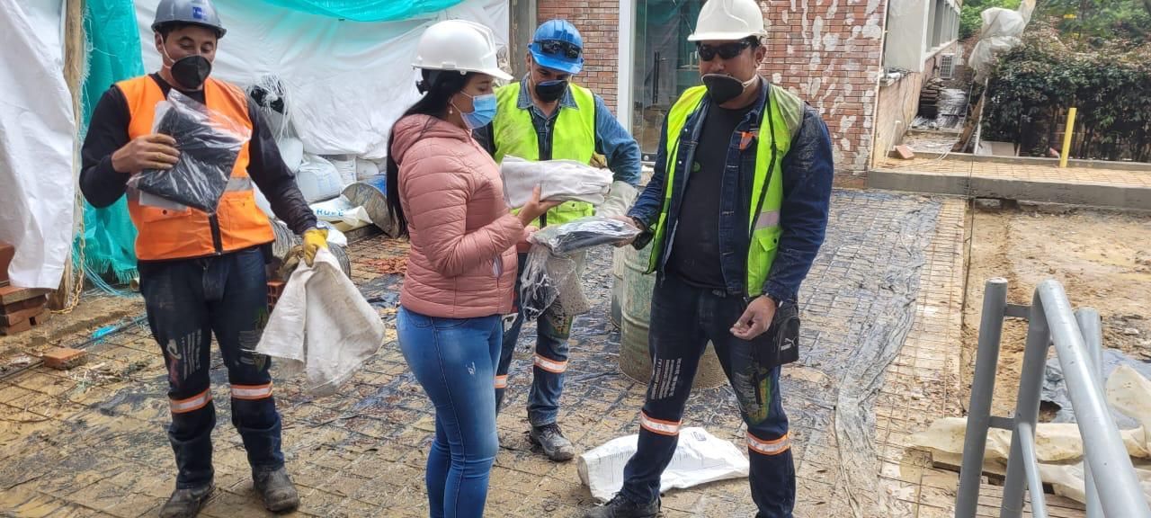 Obreros de la construcción con cascos revisan planos en un terreno fangoso con tuberías y herramientas cerca.