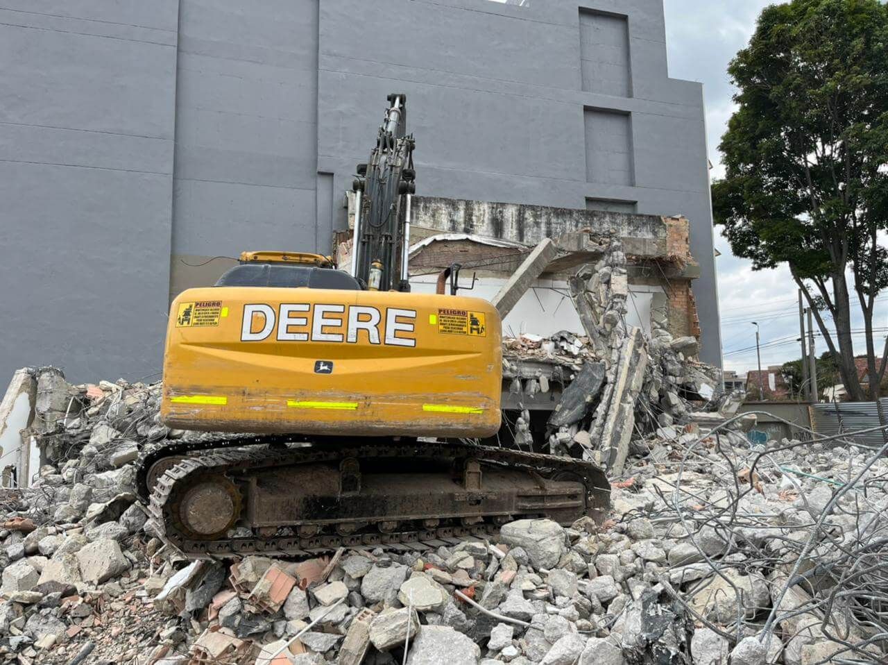 Una excavadora Deere amarilla entre los escombros, junto a un edificio gris parcialmente demolido.