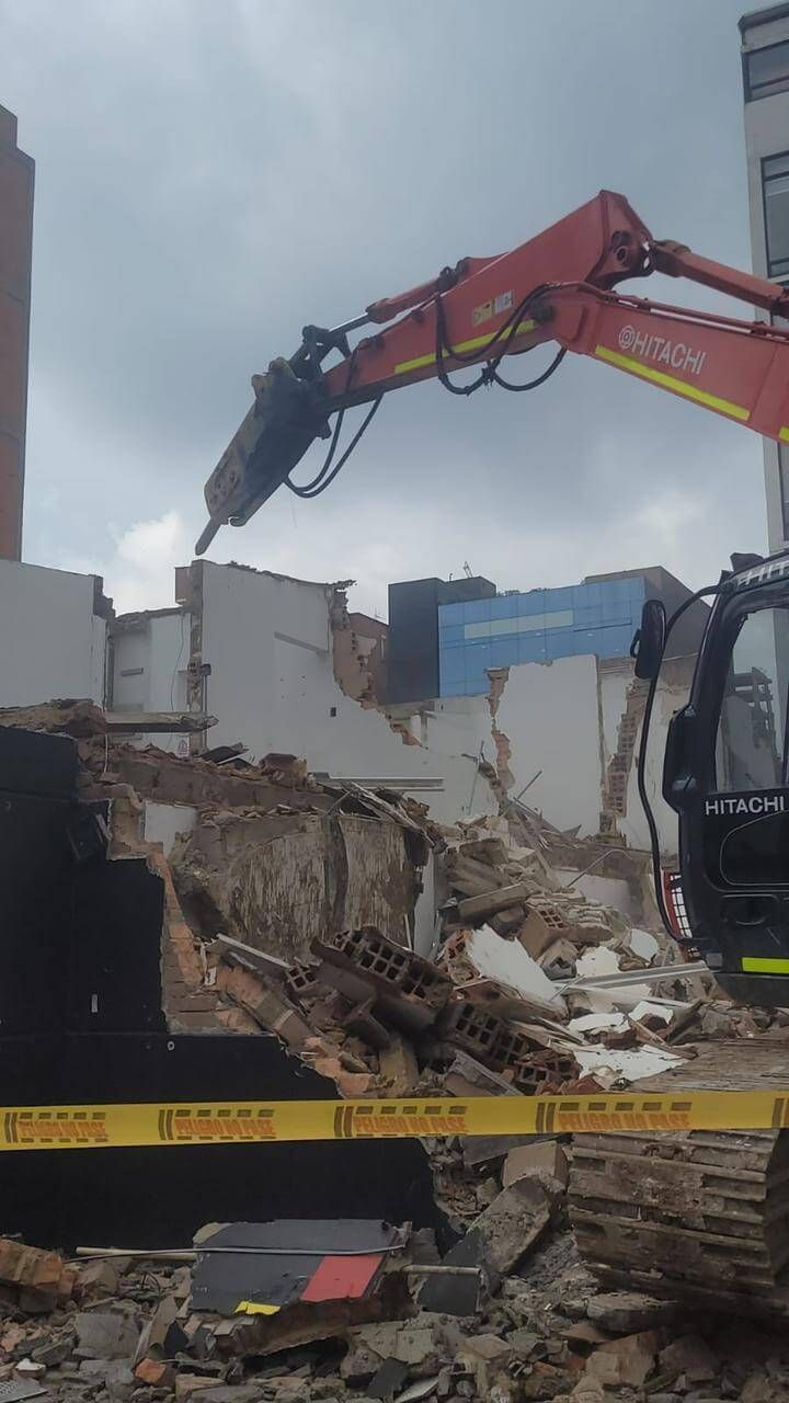 Una excavadora demuele un solar lleno de escombros detrás de una cinta amarilla de precaución.
