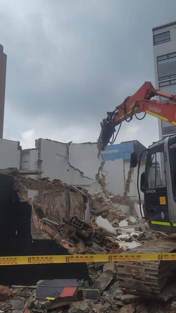 Una excavadora demuele un edificio derrumbado, con escombros, cinta de precaución y un cielo nublado.