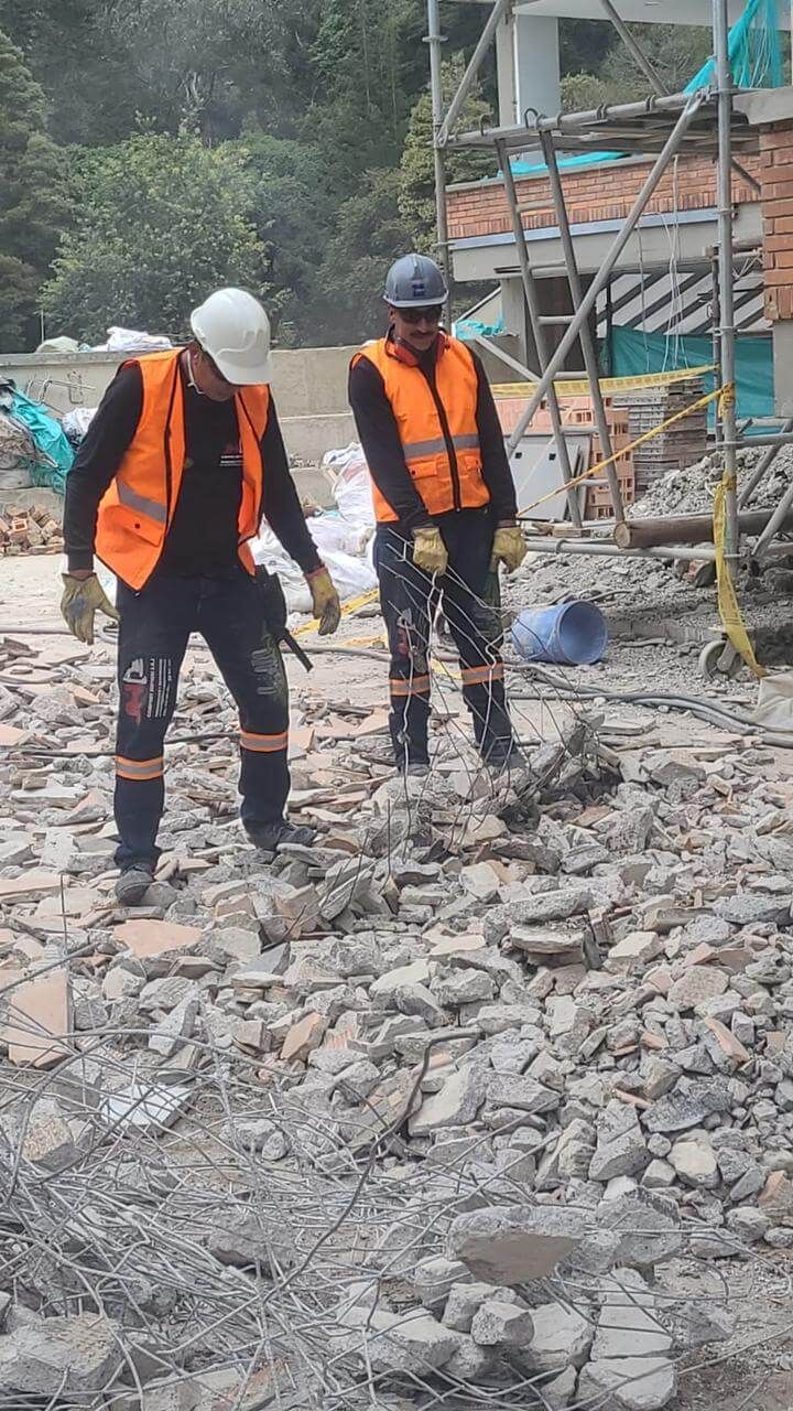 Dos obreros de la construcción, con chalecos y cascos naranjas, caminan entre los escombros de una obra en demolición.
