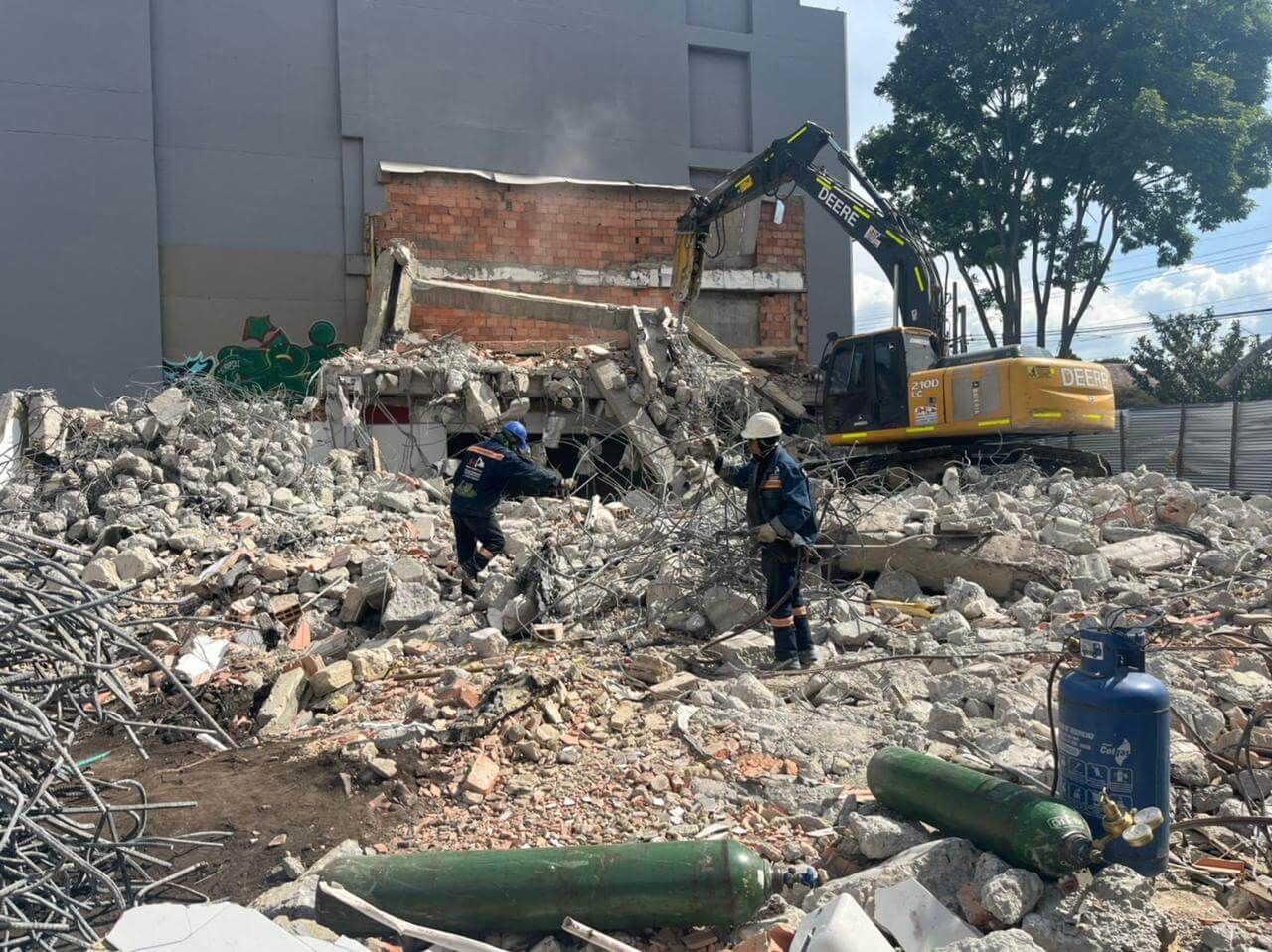 Obreros de la construcción en un solar demolido lleno de escombros junto a una excavadora y un muro de ladrillos.