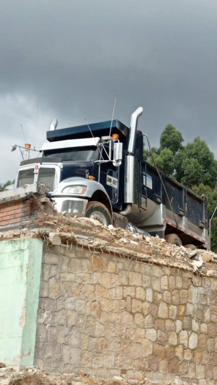 Camión volquete negro posado sobre un muro de piedra dañado bajo un cielo nublado.