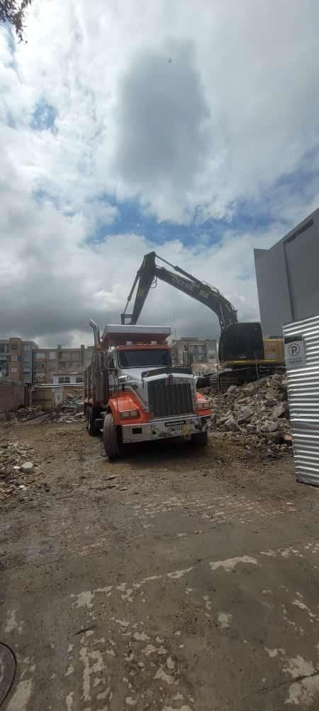 Excavadora y camión volquete en una obra de construcción o demolición llena de escombros bajo un cielo nublado.