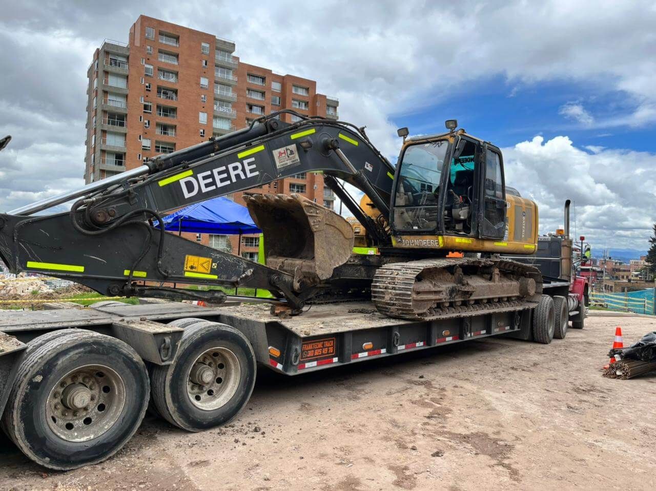 Excavadora DEERE negra sobre un remolque de plataforma en una obra de construcción con edificios de apartamentos al fondo.