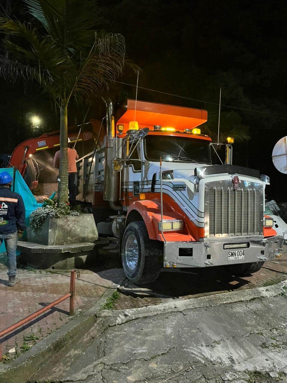 Un camión volquete naranja estacionado de noche en una obra en construcción, rodeado de palmeras y luces brillantes.
