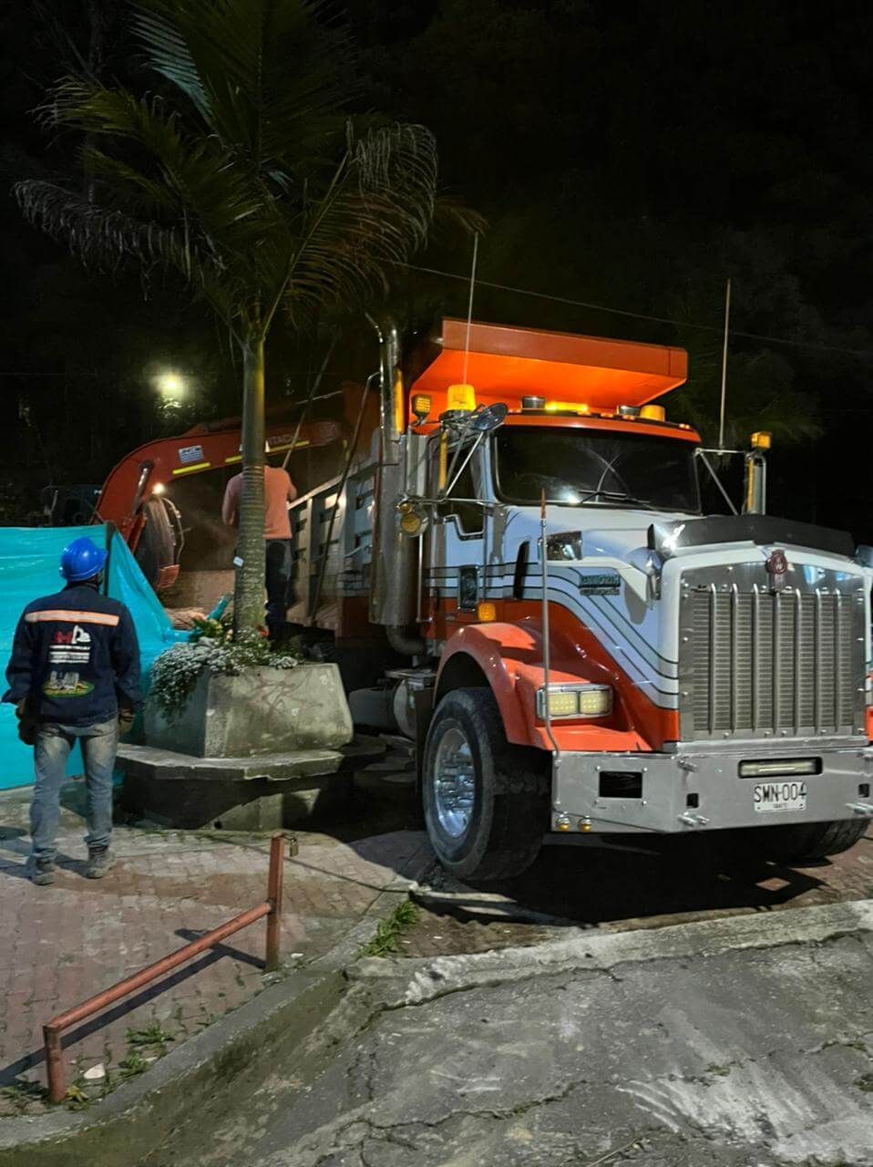 Un camión volquete naranja estacionado de noche mientras un trabajador retira escombros junto a la acera.