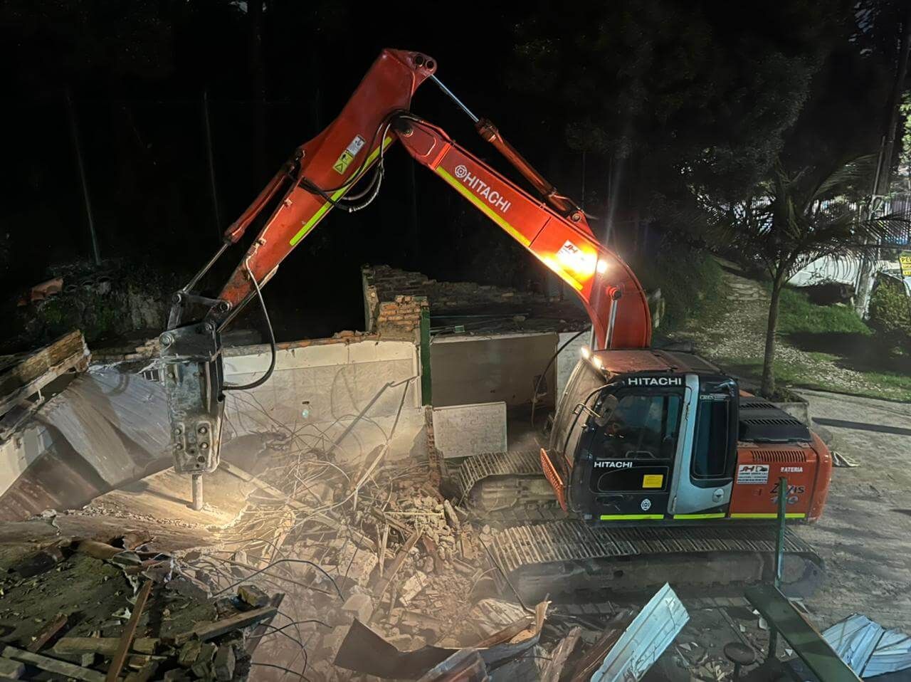 Excavadora de noche retirando escombros entre hormigón dañado y tierra, con reflectores iluminando el lugar.