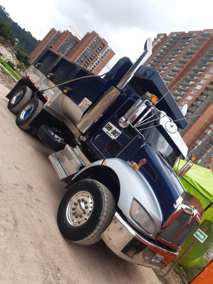 Un camión semirremolque azul y negro estacionado en ángulo en un terreno, con edificios de apartamentos al fondo.