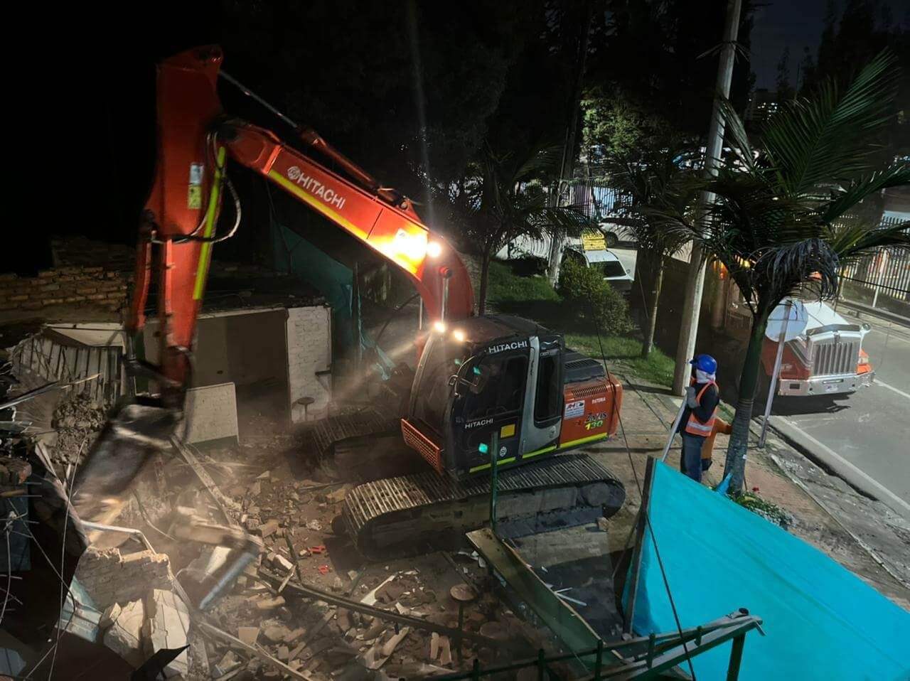 Excavadora demoliendo escombros junto a una carretera por la noche, con trabajadores y un camión cerca.