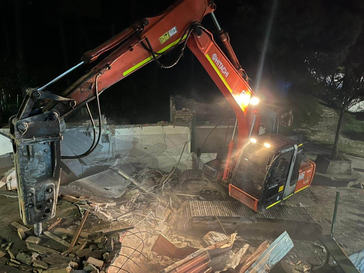 Excavadora naranja demoliendo escombros por la noche con luces de trabajo brillantes.