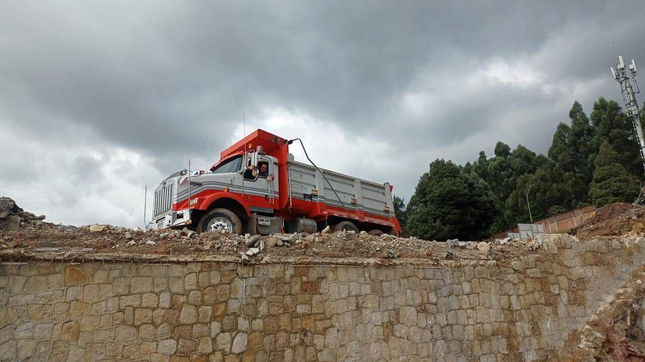 Camión volquete rojo sobre un muro de piedra bajo un cielo nublado, con árboles al fondo.
