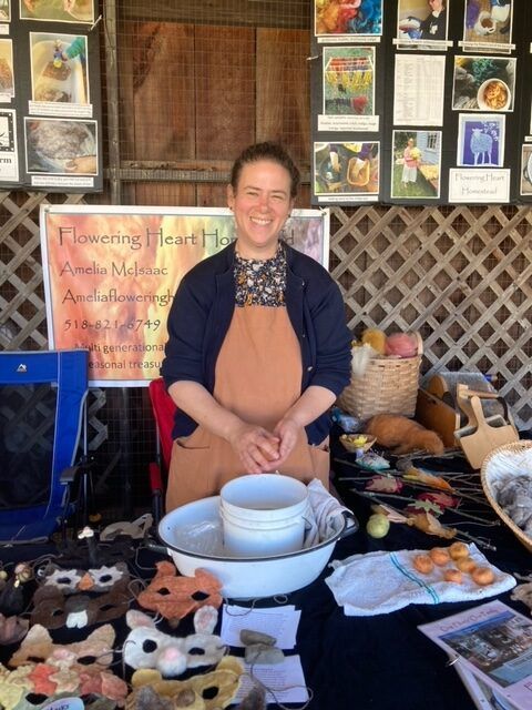 Woman at craft booth, smiling. Wearing apron, cardigan. Masks, felted wool, baskets, and sign at table.