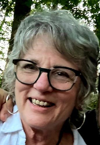 Woman with gray hair and glasses smiles, wearing a white shirt and hoop earrings.