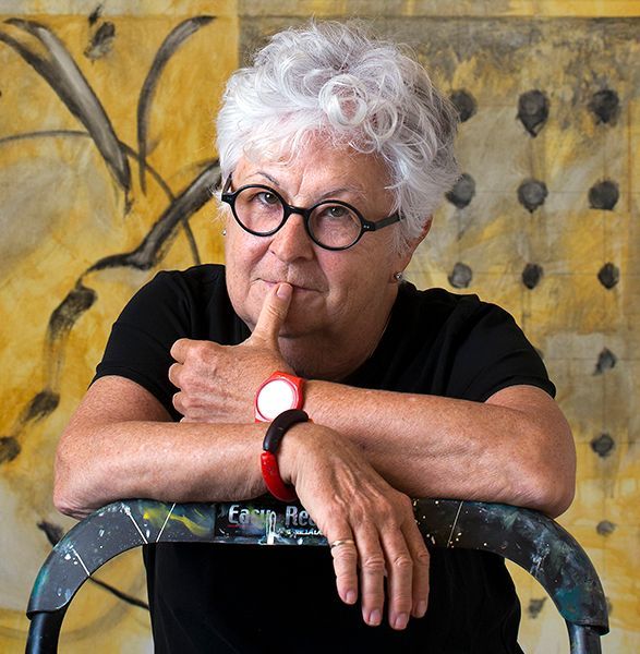 woman in black shirt, glasses, and watch, resting chin on a ladder. Yellow and black abstract art in background.