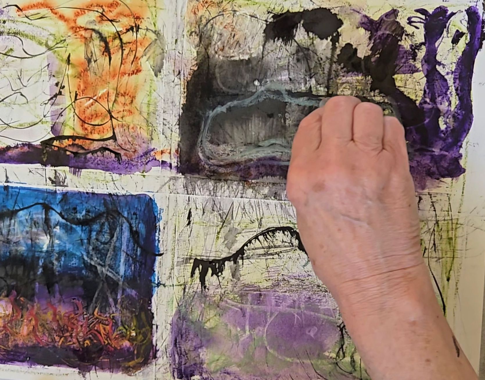 Artist's hand holding pastels, paintbrushes in a jar, with a painting of a house in the background.