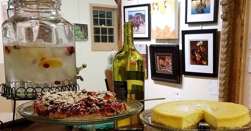 Dessert table at a gallery. Large drink dispenser, pie, cake, wine bottle, framed art.