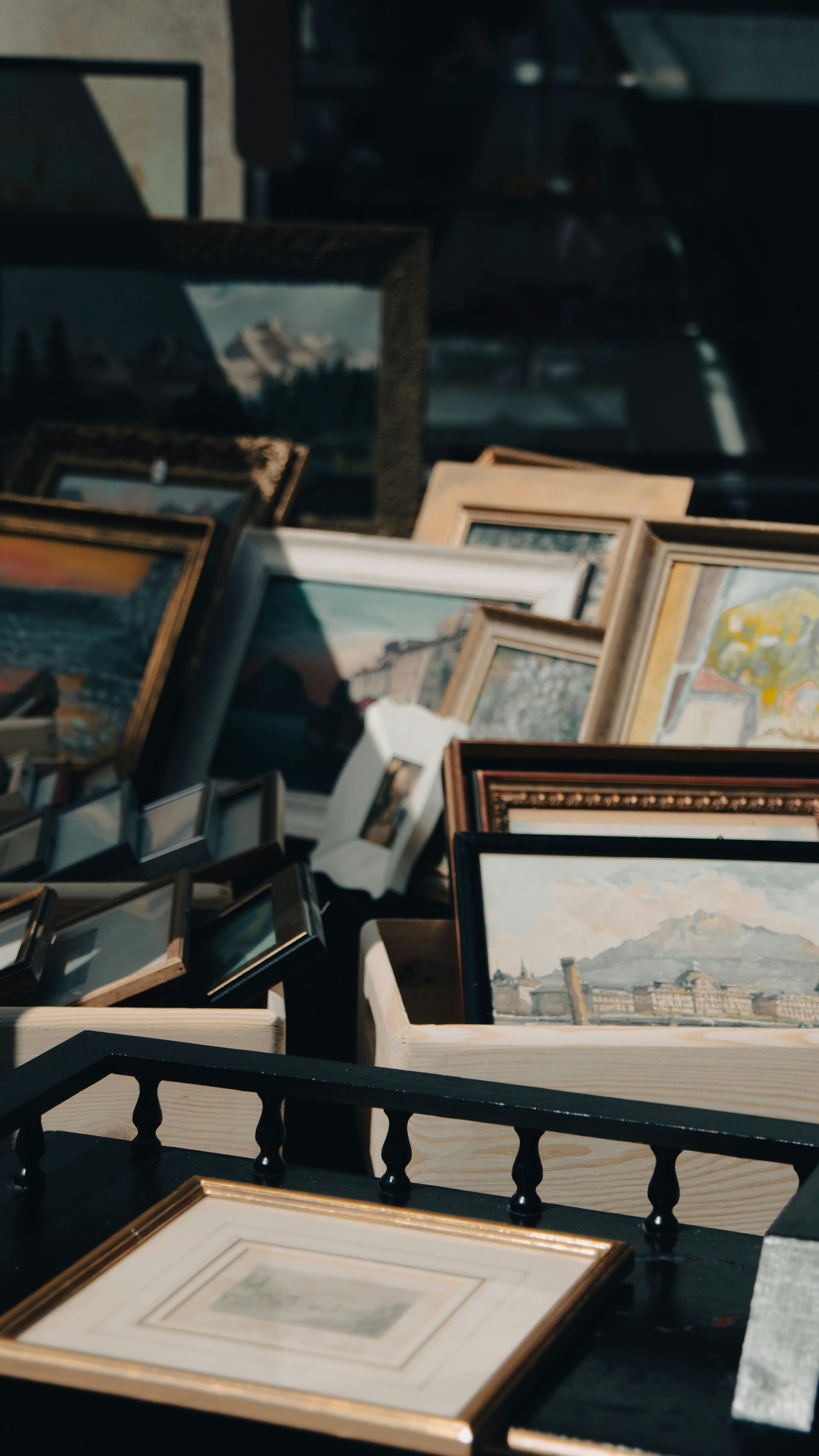 Collection of framed art, stacked and displayed on a table; sunlight and shadows visible.