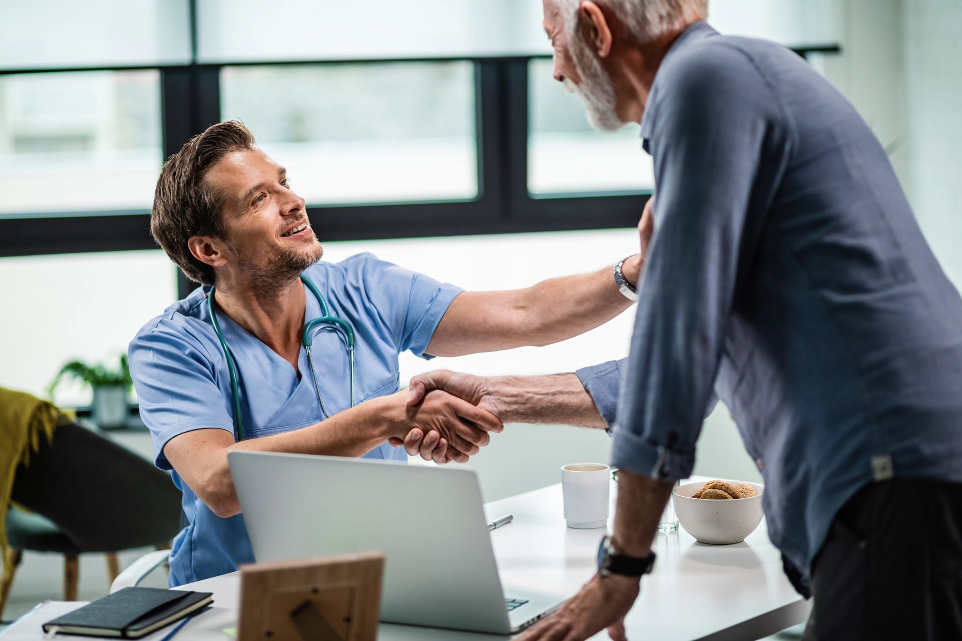 A doctor is shaking hands with an older man while sitting at a desk with a laptop.