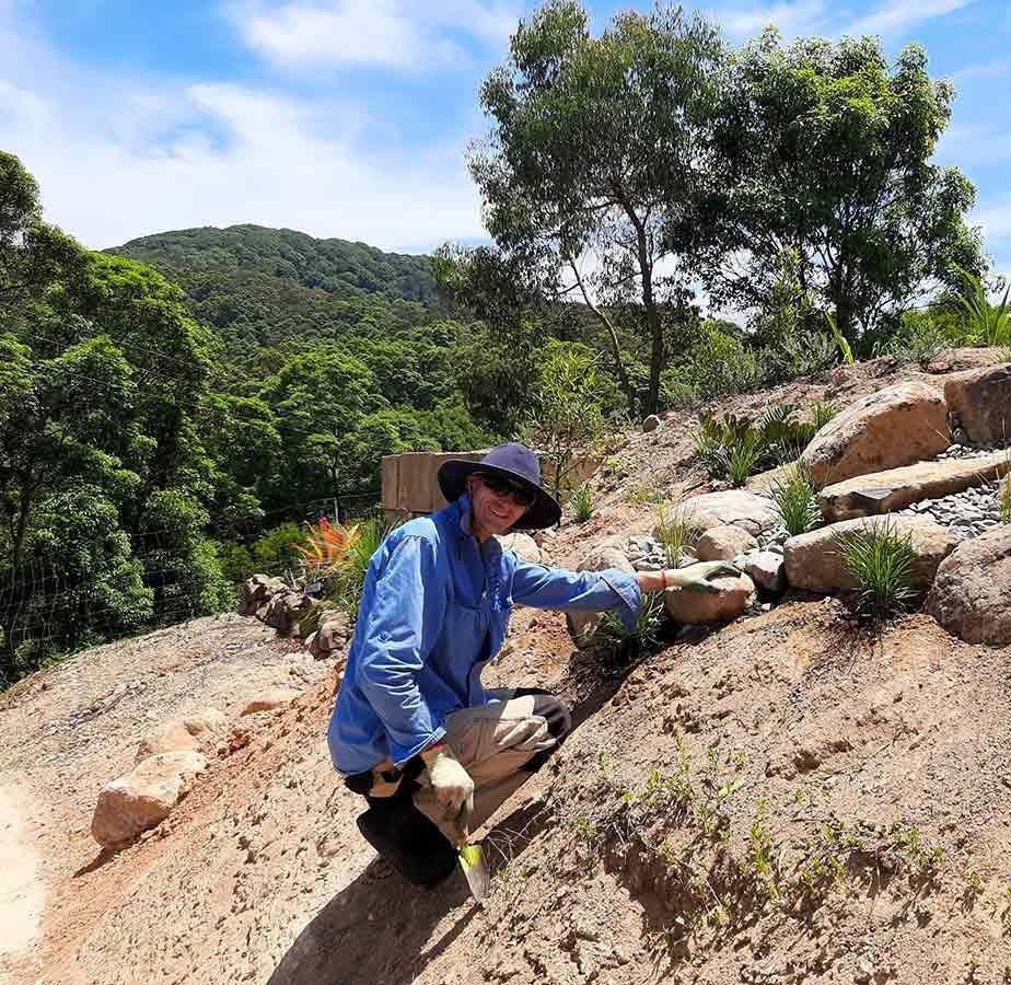 A Man in a Blue Shirt and Hat is Kneeling on a Rocky Hillside — Full Circle Landscaping in Murwillumbah, NSW