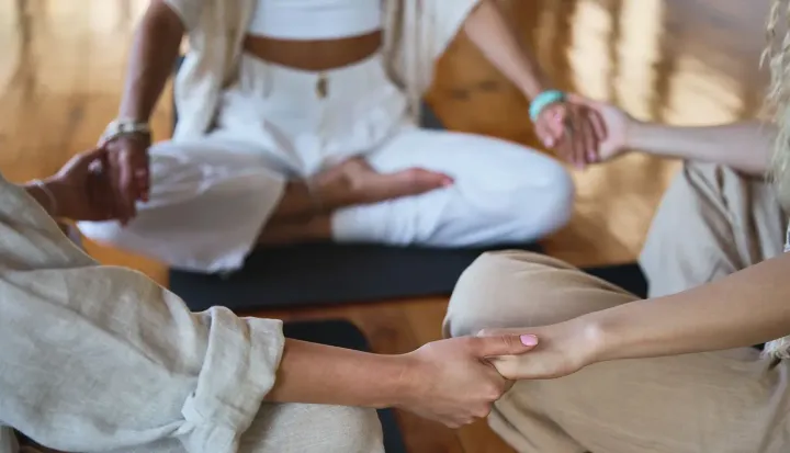 People sitting in a circle, holding hands. One person is in a lotus position, indoors, neutral tones.