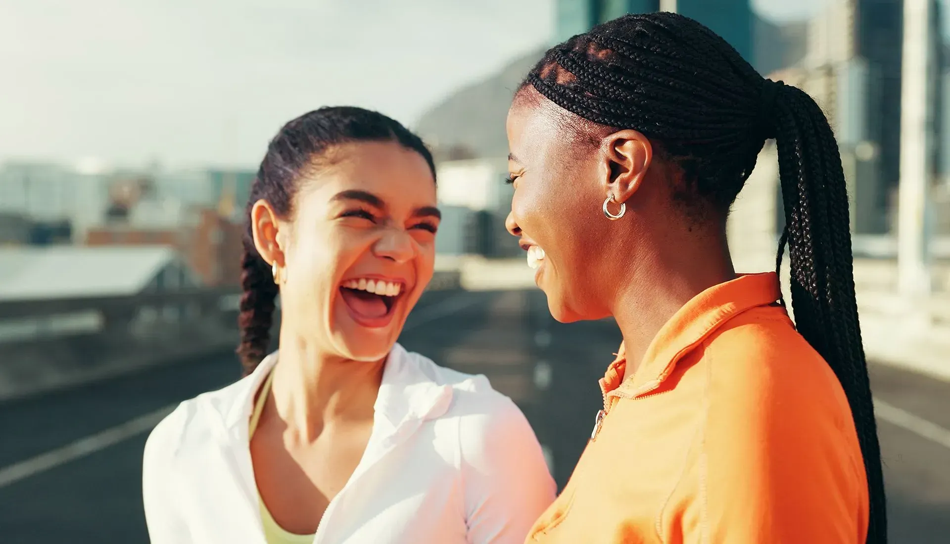 Two women laughing, outdoors. One wears orange, the other white; city background.