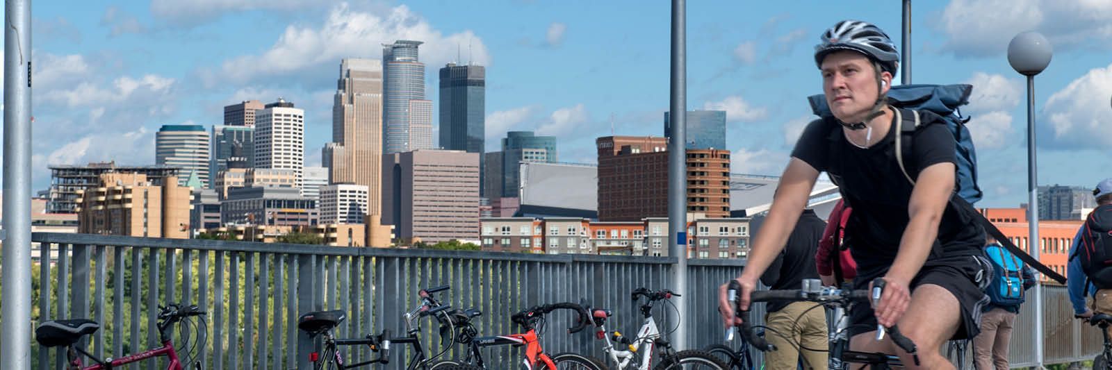 A man is riding a bike on a bridge with the Minneapolis, MN city skyline in the background.