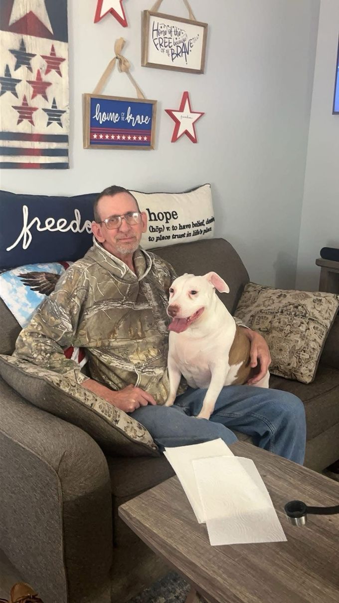Man sitting on a couch with a white dog in a decorated living room, smiling at the camera.