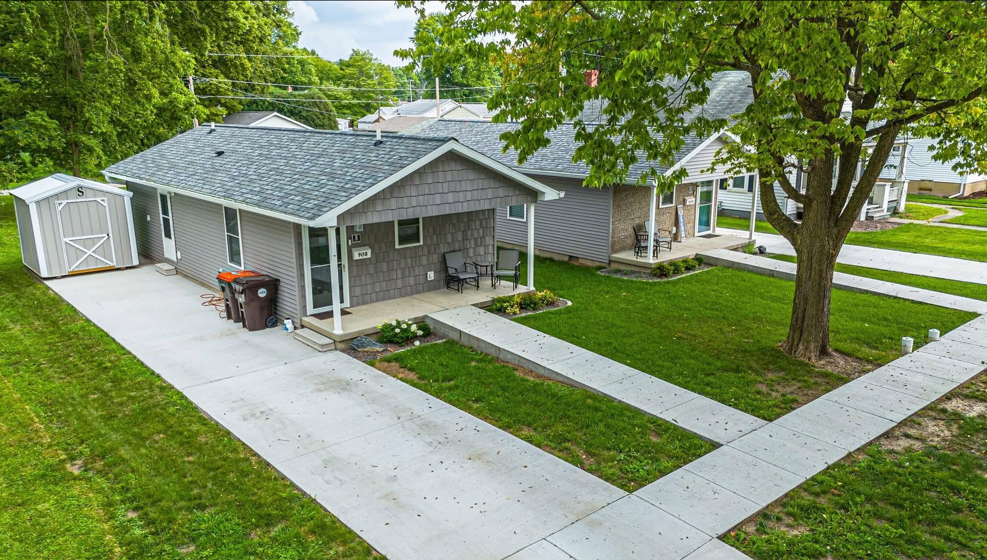 Suburban house with gray roof, front porch, concrete sidewalk, and green lawn under trees