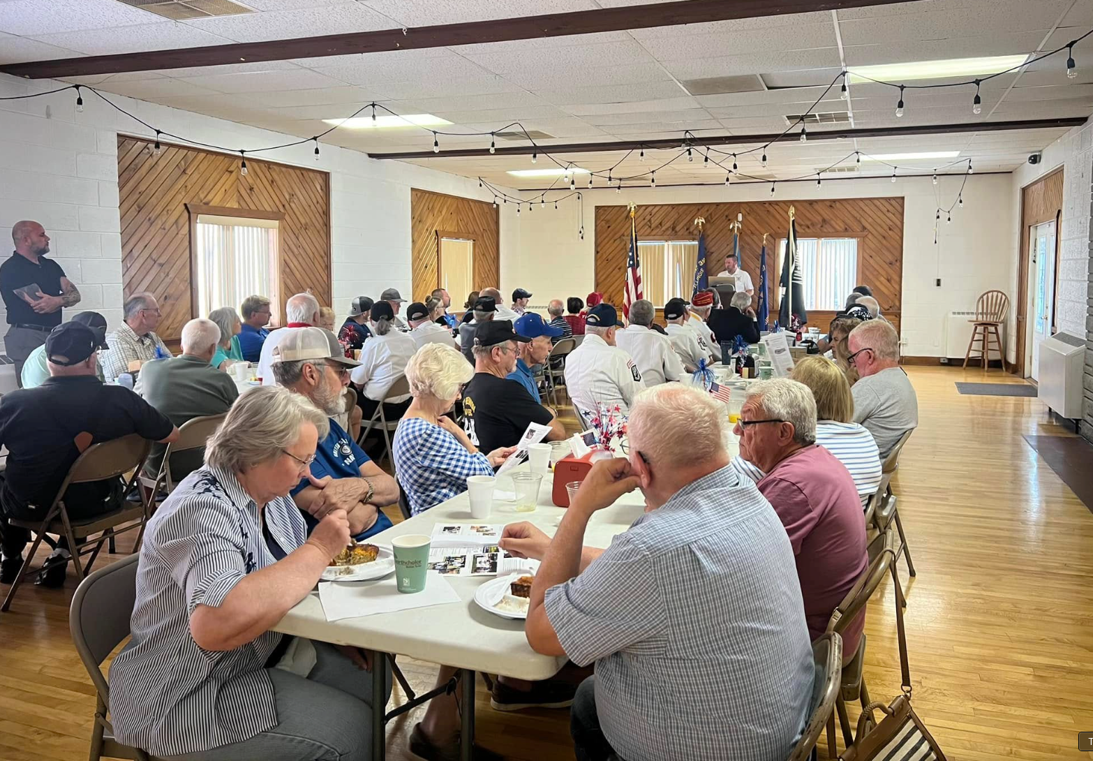 People seated at tables in a bright community hall, eating and chatting during a gathering