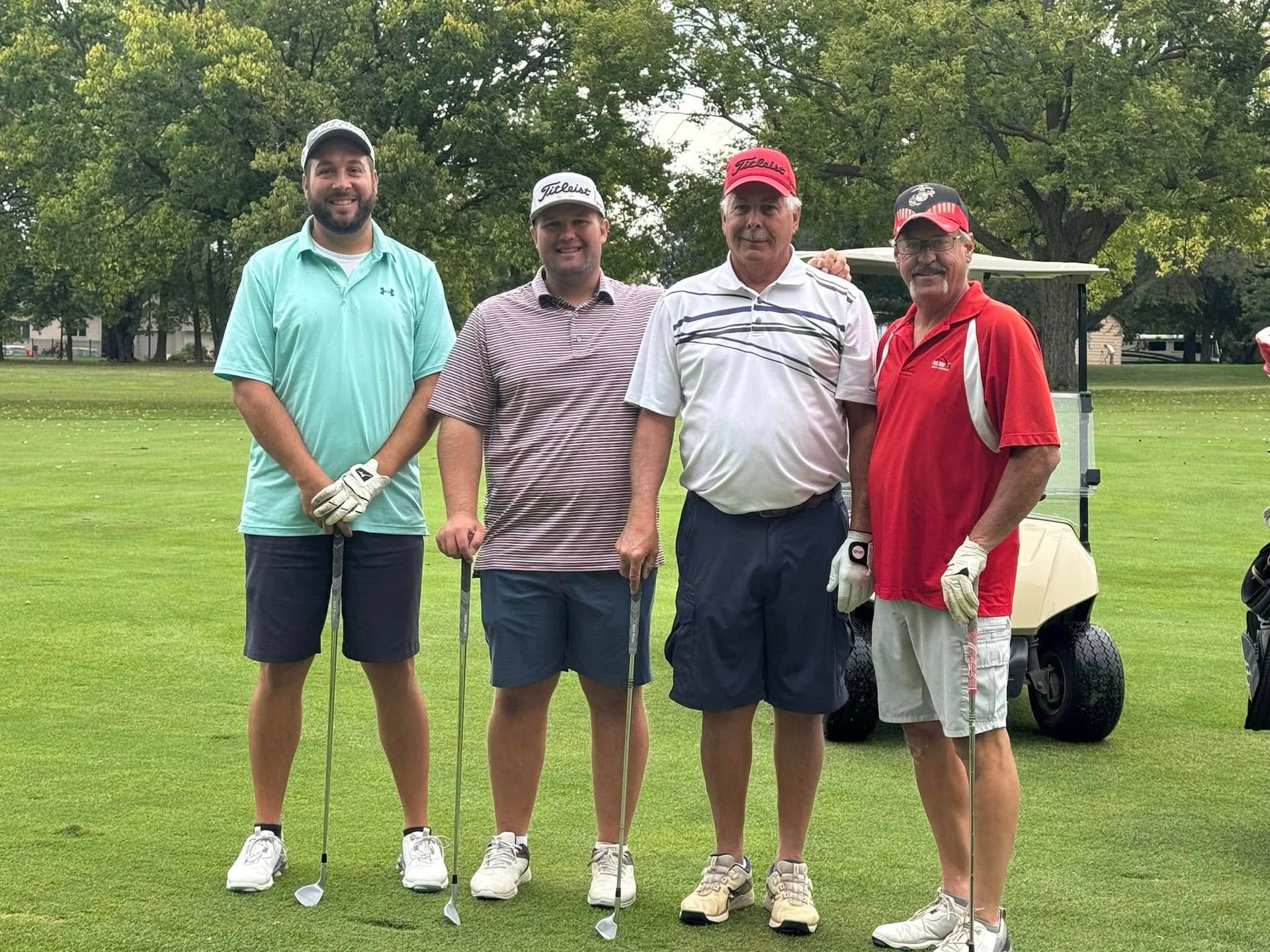 Four golfers pose on a green course, holding clubs and smiling in casual golf attire.