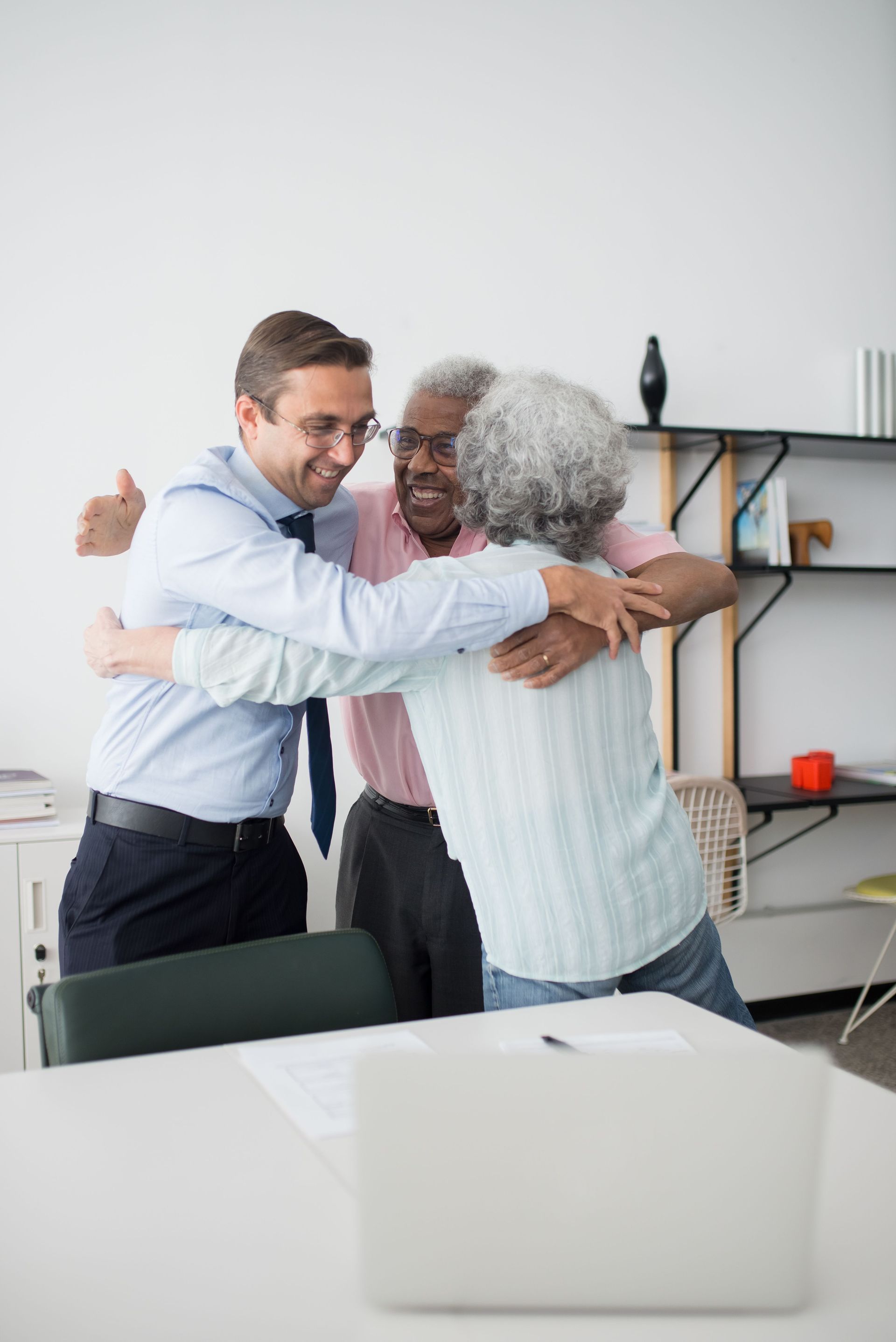 Three coworkers hugging in an office, smiling beside a table and laptop.