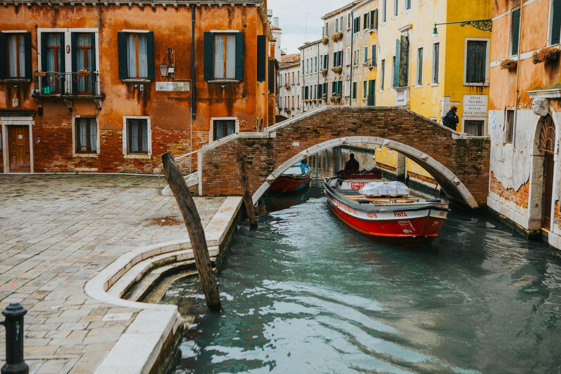 Venice canal scene with red boat under a brick bridge, buildings in warm colors frame the waterway.