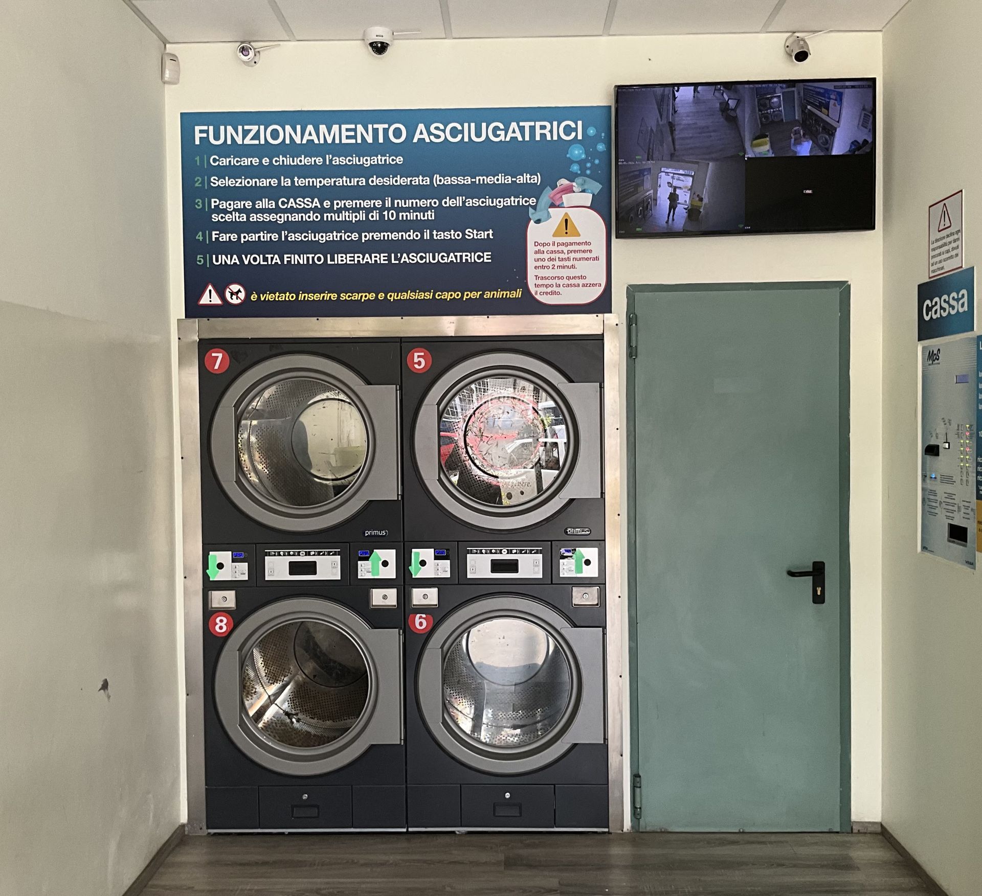 Lavanderia self-service con quattro lavatrici a carica frontale, cartello con le istruzioni e porta verde chiusa.