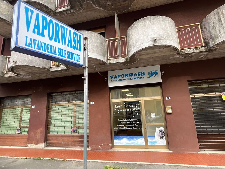 Facciata della lavanderia a gettoni Vaporwash con insegne blu e bianche e serrande metalliche chiuse in un edificio in mattoni.