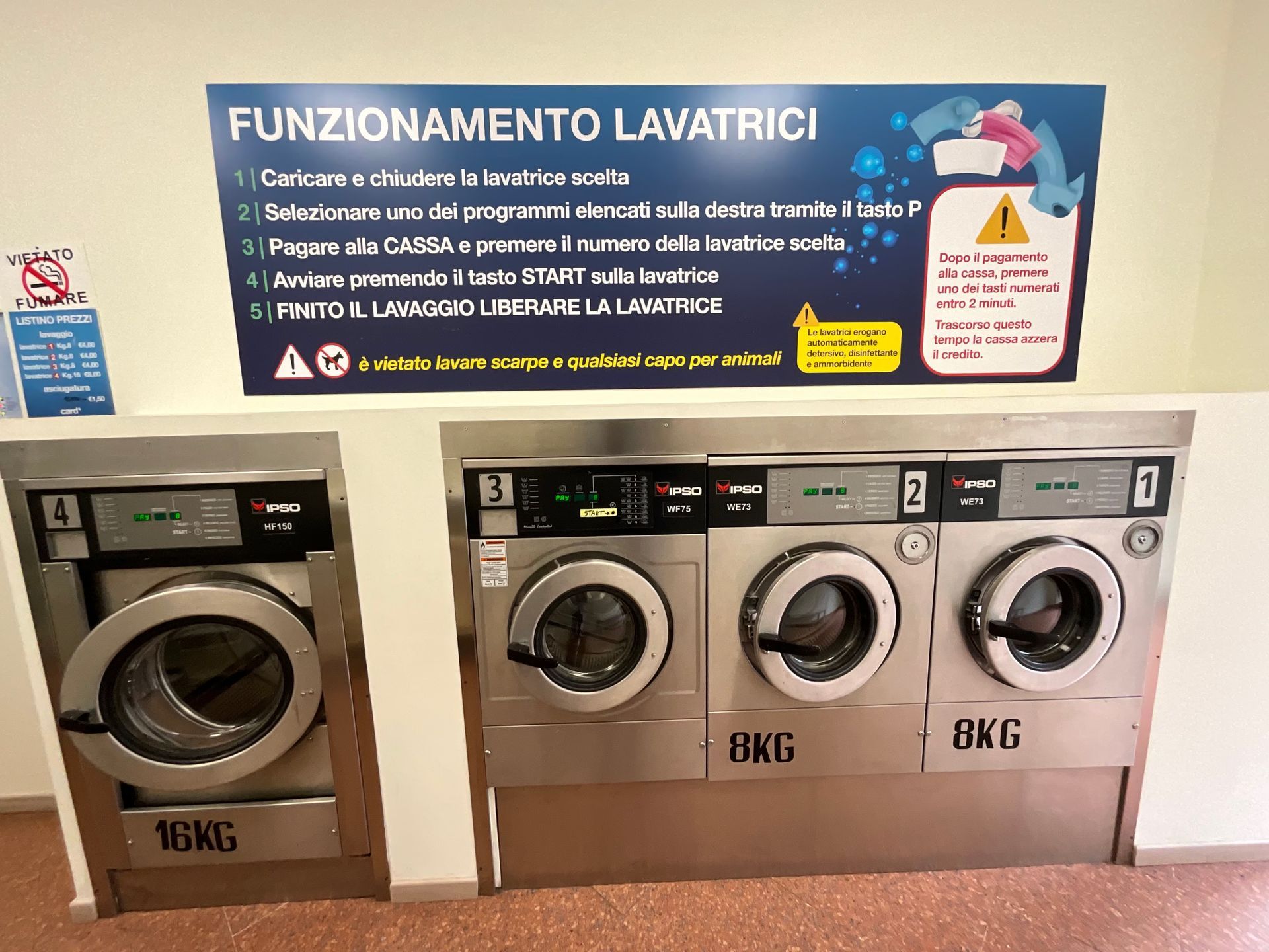 Lavanderia con quattro lavatrici a carica frontale sotto un cartello con le istruzioni in italiano.