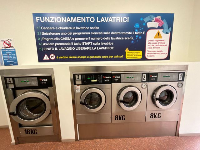 Lavanderia self-service con tre lavatrici a carica frontale e un cartello con le istruzioni in italiano sopra.